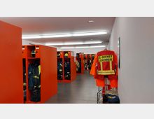 Umkleideraum einer Feuerwehrstation mit roten Spinden und darin hängender Feuerwehrschutzkleidung, Helmen und Stiefeln. Im Vordergrund hängt eine orangefarbene Jacke mit der Aufschrift „C-Dienst Luckenwalde“ an einem Kleiderständer.