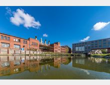Industrielle Backsteingebäude mit Schornsteinen stehen am Ufer eines Teiches, der die Bauten spiegelt, daneben ein modernes Gebäude mit Glas- und Fassadenelementen sowie einer großen Kunstinstallation. Der Himmel ist klar und blau mit wenigen Wolken.