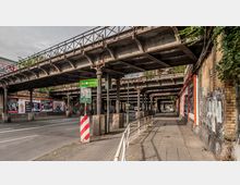 Straßenabschnitt unter einer alten, metallenen Bahnbrücke mit verzierten Säulen in Berlin-Schöneberg. Die Umgebung zeigt Werbeplakate an einer Backsteinmauer, Gehwege und Straßenschilder, umgeben von vereinzelten Pflanzen und Begrenzungspfosten.