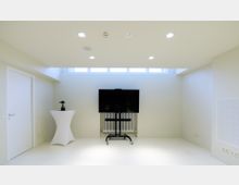 Ein minimalistisch eingerichteter Raum mit hellem Boden und weißen Wänden, ausgestattet mit einem großen schwarzen Bildschirm auf einem mobilen Gestell vor einem Heizkörper und einem hohen Stehtisch mit einer kleinen Pflanze darauf. Oben sind eingelassene Deckenleuchten und ein horizontales Fensterband, das Tageslicht hereinlässt.