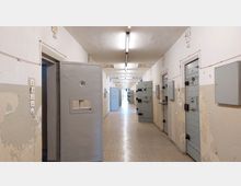 Ein langer, kahler Gefängnisflur mit weißen Wänden und abblätternder Farbe, flankiert von massiven grauen Zellentüren mit kleinen vergitterten Sichtfenstern und schweren Schlössern. Die Decke ist mit einfachen Leuchtstofflampen beleuchtet, und der Boden ist mit einem gemusterten Belag ausgelegt.