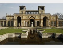 Neoklassizistisches Gebäude mit gelber Fassade und symmetrischen Arkaden, flankiert von Türmen mit offenen Durchgängen. Im Vordergrund ein symmetrisch gestalteter Garten mit einem Brunnen und einer Statue auf einem Sockel.