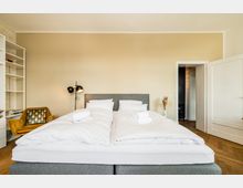 Helles Schlafzimmer mit zwei zusammengestellten Einzelbetten mit weißer Bettwäsche und grauem Kopfteil. Links stehen ein offenes Regal, ein gepolsterter Sessel und eine schwarze Stehlampe; rechts führt eine offene Tür in einen Flur, der Boden ist aus Holzparkett.