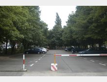 Parkplatz mit mehreren Autos, umgeben von dicht bewachsenen Bäumen. Im Vordergrund befindet sich eine Schranke, die den Zugang zum Parkplatz kontrolliert.