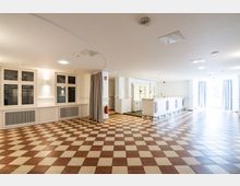 Heller Innenraum mit einem rot-weiß karierten Boden, cremefarbenen Wänden und großen Fenstern. Auf der rechten Seite befindet sich ein langer Tresen in weißer Ausführung, dahinter ein Bereich mit Regalen und Lampen.