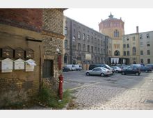 Ein Innenhof mit Kopfsteinpflaster, umgeben von historischen Gebäuden aus Backstein, darunter ein Turm mit dekorativen Fensterelementen. Im Vordergrund befinden sich Briefkästen an einer Mauer sowie ein roter Hydrant, während der Hof als Parkplatz mit mehreren geparkten Fahrzeugen genutzt wird.