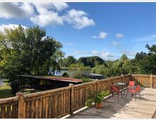 Holzterrasse mit Geländer, darauf ein runder Tisch mit zwei roten Stühlen und einem Pflanzentopf; im Hintergrund Bäume, ein kleiner See und ein Pavillon mit rot gedecktem Dach unter blauem Himmel.