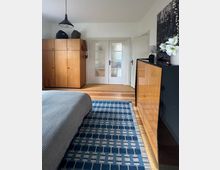 Schlafzimmer mit Holzboden und blau gemustertem Teppich, links ein Bett sowie ein großer Holzkleiderschrank unter einer Hängelampe. Hinten stehen weiße Doppeltüren mit Milchglas, rechts eine Kommode und eine offene Tür zu einem Nebenraum.