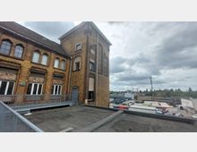 Ziegelsteingebäude mit industriellem Charakter und einem Turm, auf dessen Fassade bogenförmige Fenster und Verzierungen zu sehen sind. Im Hintergrund ein Industriegebiet mit Containern, Fahrzeugen und einem Sendemast unter bewölktem Himmel.