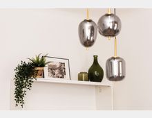 Deutsche Beschreibung:  
Nahaufnahme eines weißen Regals mit dekorativen Elementen, darunter eine hängende grüne Pflanze in einem Korb, ein gerahmtes Schwarz-Weiß-Foto und grüne Glasvasen. Im Vordergrund hängen drei silberne, glänzende Lampen mit goldenen Halterungen.