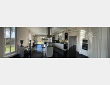 Moderne Küche mit weißen Schränken und Hochglanzarbeitsflächen, einer zentralen Kochinsel mit Barhockern und einem Edelstahl-Dunstabzug. Große Fenster und eine Glastür bieten Blick auf einen Garten mit Rasen und Bäumen.