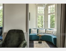 Wohnzimmerbereich mit einer gebogenen, türkisfarbenen Couch vor einem Erkerfenster mit Sprossen. Im Vordergrund ein dunkelgrüner Sessel, ein kleiner Beistelltisch mit Glas und Metall sowie ein heller Teppich. Außen sind grüne Bäume sichtbar.