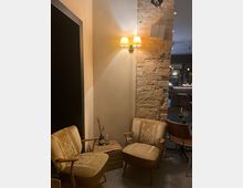 Gemütliche Sitzecke in einem Innenraum mit zwei beigen Polstersesseln und einem kleinen Holztisch, dekoriert mit einer Vase. Eine Backsteinmauer mit einer angeschalteten Wandlampe bildet die Hintergrundkulisse.