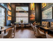 Schick eingerichtetes Café mit dunklen Wänden, großen Fenstern und Kunstwerken an den Wänden. Der Raum verfügt über moderne, gepolsterte Stühle, Holzböden und Tische, die mit minimalistischen Dekorationen ausgestattet sind.