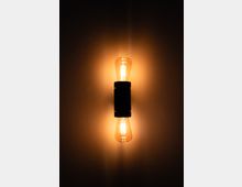 Eine Wandlampe mit zwei Glühbirnen, die nach oben und unten gerichtet sind, leuchtet warmes, gelbliches Licht auf eine glatte, dunkle Wand.