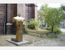 Bronzeskulptur einer kopflosen, torsoartigen Figur auf einem Sockel vor einer Fassade aus rotbraunem Backstein. Im Hintergrund ein kleiner, begrünten Gartenbereich mit Bäumen und umliegenden Gebäuden.