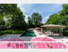 Grüner Garten mit einem rechteckigen Swimmingpool, umgeben von einer gepflasterten Terrasse mit rosa Sonnenschirmen und einem modernen Pavillon mit Glaswänden. Im Hintergrund stehen hohe Bäume, die die Umgebung abschirmen.