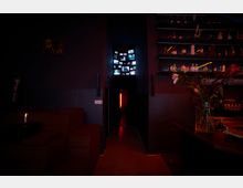 Dunkles, atmosphärisches Bar-Interieur mit einer schmalen Türöffnung, die zu einem rot beleuchteten Korridor führt. Im oberen Bereich der Tür befinden sich mehrere kleine Bildschirme mit verschiedenen Videos, während auf der rechten Seite Regale mit Flaschen und Blumen in einer Vase auf einem Tresen zu sehen sind.