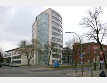 Ein modernes Bürogebäude mit abgerundeter Glasfassade an der Ecke einer Straßenkreuzung. Im Vordergrund sind Bäume ohne Laub, geparkte Autos und Verkehrsschilder sichtbar, im Hintergrund befinden sich ein rotes Backsteingebäude und weitere moderne Gebäude.