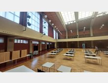 Großer Veranstaltungssaal mit Holzboden, großen Fenstern und einer hohen Decke mit Oberlichtern. Im Raum stehen vereinzelte Tische und Stühle in Reihen, an den Seiten befinden sich Vorhänge und Beleuchtungseinrichtungen.
