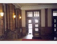 Eingangsbereich eines historischen Gebäudes mit dekorativen, braunen Steinsäulen, verzierten Glastüren und Decken mit klassischen Ornamenten. Wandleuchten beleuchten den Raum, und eine Treppe führt nach oben.