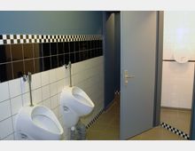 Öffentliche Herrentoilette mit zwei Urinalen an einer Wand mit schwarz-weißen Fliesen. Im Hintergrund befindet sich eine geöffnete Tür, die zu einem weiteren Bereich führt.