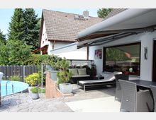 Terrasse eines Einfamilienhauses mit Pflastersteinboden, überdacht durch eine Markise. Zu sehen sind eine Sitzecke mit Sofa und Liege, Pflanzen in Töpfen, ein Esstisch mit Stühlen sowie ein kleiner Pool im Hintergrund, umgeben von einer Hecke und einem Zaun.