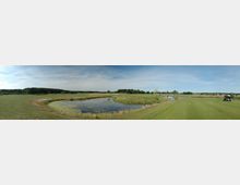 Weitläufige Golfplatzlandschaft mit gepflegten grünen Fairways, einem kleinen Teich in der Mitte und umgeben von Wiesen und Bäumen. Im Hintergrund sind ein Waldgebiet und einige Gebäude am Horizont zu sehen, während ein Golfcart am rechten Bildrand geparkt ist.