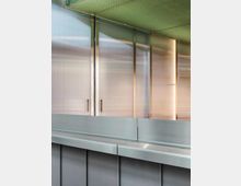 Eine Innenansicht mit Metall- und Glasoberflächen, darunter eine graue Theke und transparente Wandpaneele mit Metallrahmen. Die Decke ist mit einem grünen, gitterartigen Muster gestaltet.