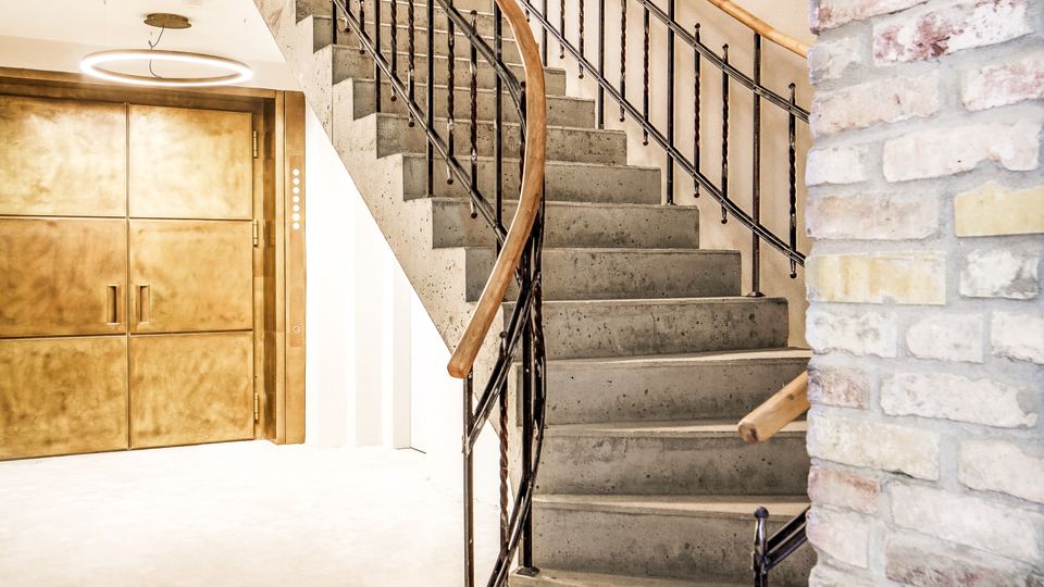 Innenansicht eines Treppenhauses mit breiter Betontreppe und schwarzem Metallgeländer mit Holzhandlauf, die in einem Bogen nach oben führt. Helle Wände, eine sichtbare Backsteinwand im Vordergrund und doppelflügelige Holztüren im Erdgeschoss prägen den Raum.