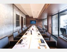 Langer, modern eingerichteter Konferenzraum mit einem gedeckten Tisch, umgeben von schwarzen Ledersesseln. Links eine Steinwand mit Fenstern, rechts eine Glasfront mit Blick nach draußen; im Hintergrund ein Bildschirm auf einer Holzvertäfelung.