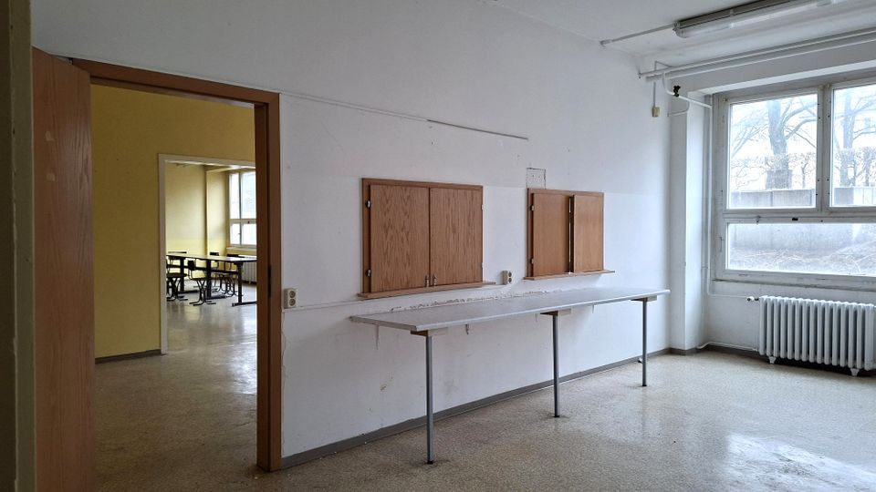 Leerer Innenraum in einem älteren Gebäude mit weißen Wänden, einer langen schmalen Ablage auf Metallbeinen und zwei an der Wand montierten Holzschränken. Rechts befindet sich ein großes Fenster mit Heizkörper darunter, links ein offener Türrahmen mit Blick in einen Nebenraum mit Tischen.