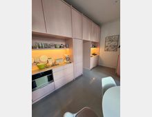 Moderne Küche mit pastellrosafarbenen Einbauschränken und offenen Regalfächern, die beleuchtet sind. Im Vordergrund ein runder weißer Tisch mit Stühlen, der Boden besteht aus glattem grauem Material.