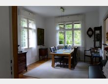 Essbereich in einem hellen Raum mit großen Fenstern, einem hellbraunen Holztisch und passenden Stühlen. Im Raum befinden sich ein Teppich auf Holzboden, eine antike Wanduhr und ein Sideboard, während Blick nach draußen viel Grün zeigt.