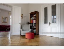 Wohnraum mit hellem Holzboden, einem grauen Sessel, einem roten Hocker und einem Holzregal voller Bücher. Links ist eine offene Tür zu einem weiteren Raum mit rotem Sofa, die weißen Doppeltüren rechts haben Glaseinsätze.