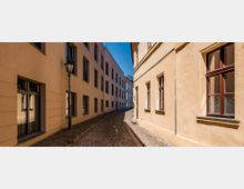 Schmale, gepflasterte Gasse zwischen hellen Gebäuden mit rechteckigen Fenstern; auf der linken Seite steht eine Straßenlaterne, und im Hintergrund scheint blauer Himmel durch.