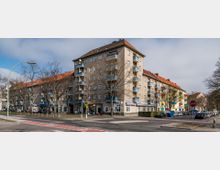 Wohnhauskomplex an einer Straßenecke mit vierstöckigen Gebäuden, hellbrauner Fassade und blauen Balkonbrüstungen. Im Erdgeschoss befinden sich Geschäfte und Restaurants; die Straße ist von parkenden Autos und kahlen Bäumen gesäumt.