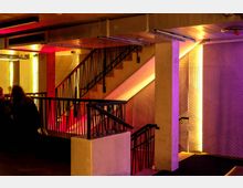 Innenansicht eines modernen Treppenhauses mit Betonwänden und schwarzen Geländern. Die Wände sind mit vertikalem Holzmuster verkleidet und durch gelb-rötliche sowie violette Lichtakzente beleuchtet.