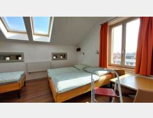 Ein schlichtes Schlafzimmer mit weiß gestrichenen Wänden, zwei Einzelbetten mit grün gemusterter Bettwäsche, einem Holzboden und einem Tisch mit Stuhl. Das Zimmer hat ein großes Fenster mit orangefarbenen Vorhängen sowie zwei Dachfenster, die viel Tageslicht hereinlassen.