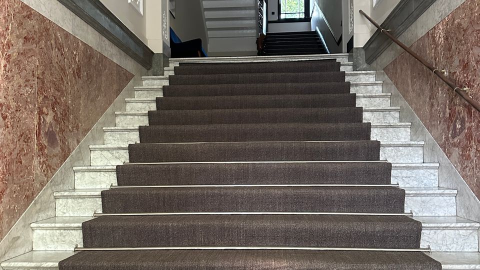 Innenansicht eines historischen Treppenhauses mit breiter Marmortreppe und dunklem Teppichläufer, flankiert von marmorverkleideten Wänden und Handläufen. Oben sind Stuckdecken und hängende Kugellampen zu sehen, am Ende des Flurs ein großes Fenster mit farbigem Glas.
