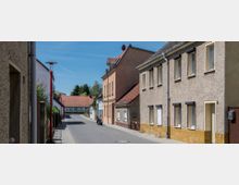 Straßenansicht mit Wohnhäusern in einer ruhigen, ländlichen Siedlung; die Gebäude haben Putzfassaden mit vereinzelten Fenstern, teilweise mit geschlossenen Rollläden. Im Hintergrund befinden sich weitere Häuser mit roten Ziegeldächern sowie Bäume.