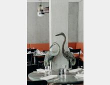 Eine moderne Restaurantinneneinrichtung mit Tischen, die mit Gläsern und weiß gefalteten Servietten gedeckt sind. Im Vordergrund stehen zwei dekorative Vogelstatuen aus Metall vor einer Betonstütze, mit orangen Sitzbänken und grauen Wänden im Hintergrund.