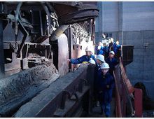 Eine Gruppe von Menschen in Schutzhelmen und Arbeitskleidung besichtigt das Innere einer industriellen Anlage mit metallischen Strukturen, Rohren und Maschinen. Im Hintergrund sind graue Fabrikwände sichtbar.