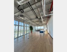 Open-plan modern office lounge with light wood floors, floor-to-ceiling windows along one wall, and an exposed industrial ceiling with visible ducts, pipes, and wiring. The space includes a potted plant, seating area, and a foosball table, with city views through the windows.