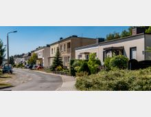 Siedlungsstraße mit modernen Reihenhäusern, umgeben von gepflegten Gärten und vereinzelten Bäumen; auf der Straße sind parkende Autos zu sehen, daneben führt ein schmaler Gehweg entlang.