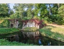 Steinerne Grotte mit gewölbtem Eingangsbereich, umgeben von roten und grünen Kletterpflanzen, steht an einem kleinen Teich inmitten eines bewaldeten Parks. Die Umgebung ist von üppigem Grün und reflektierendem Wasser geprägt.