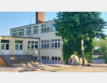 Zweistöckiges Schulgebäude mit gelber Fassade, zahlreichen weißen Fenstern und einem hohen Backsteinkamin. Im Vordergrund befinden sich eine breite Treppe mit Geländern, ein großer Baum, eine teilweise bewachsene Wand und ein Basketballkorb im Hintergrund.