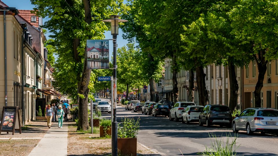 Baumbestandene Stadtstraße mit Gehweg und parkenden Autos entlang niedriger, historischer Gebäude mit Ladenfronten und Cafébestuhlung. In der Straßenmitte stehen eine Laterne mit Banner und ein Schild „Kirchplatz“, die Straße führt geradeaus in ein dicht begrüntes Viertel.