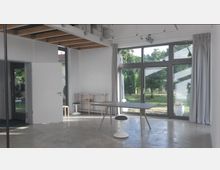 Modernes Zimmer mit großen Glasfenstern, die einen Blick auf einen grünen Garten bieten. Der Raum hat eine minimalistische Einrichtung mit einem schlichten Tisch, Hockern und einem Regal, während die Decke sichtbare Holzbalken aufweist.