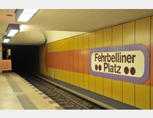 U-Bahnhof mit gelb-orangefarbenen Wandpaneelen, großem lila-umrandeten Stationsschild mit der Aufschrift „Fehrbelliner Platz“ und Schienen, die in einen dunklen Tunnel führen. Deckenleuchten beleuchten den Bereich.